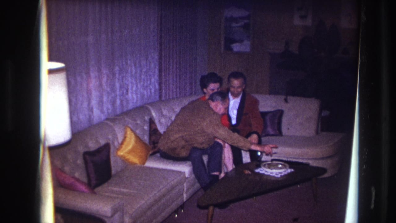 In a 1967 Ottawa home-movie frame, three adults sit closely on a sectional couch as a man leans toward a dark coffee table.