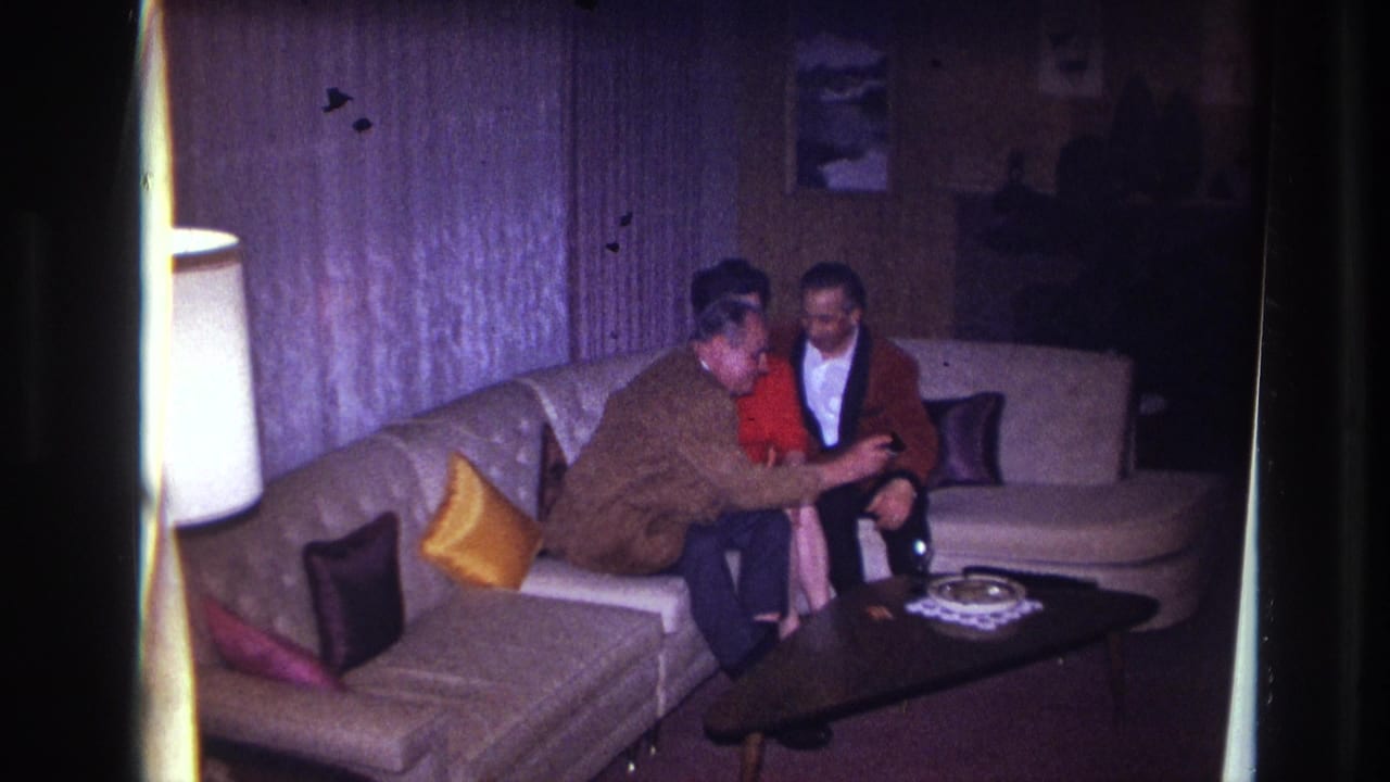 Three adults sit together on a couch, with one man leaning forward near a coffee table and lamp.