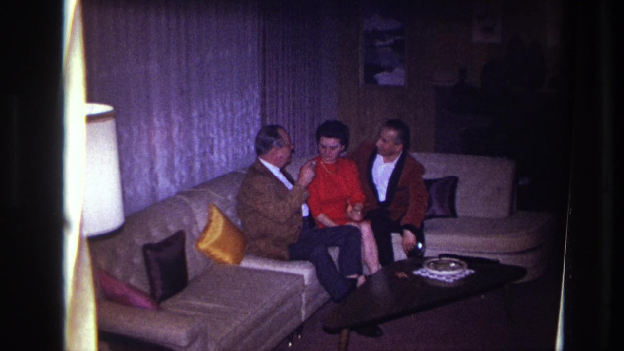 In a 1967 Ottawa home-movie frame, three adults sit close together on a pale sectional sofa in a dim living room.