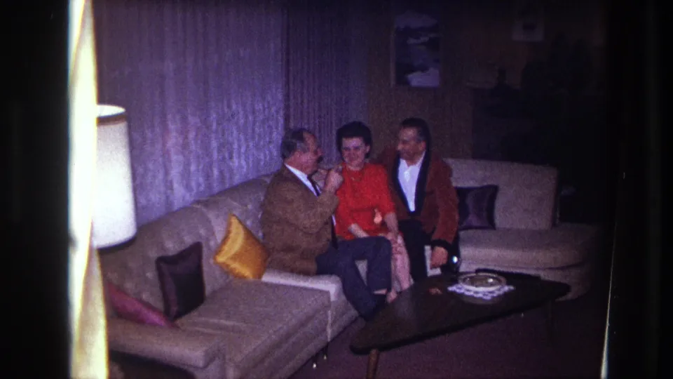 The home-movie frame shows three adults gathered on a pale sofa, with colorful pillows, a table ashtray, and curtains behind them.