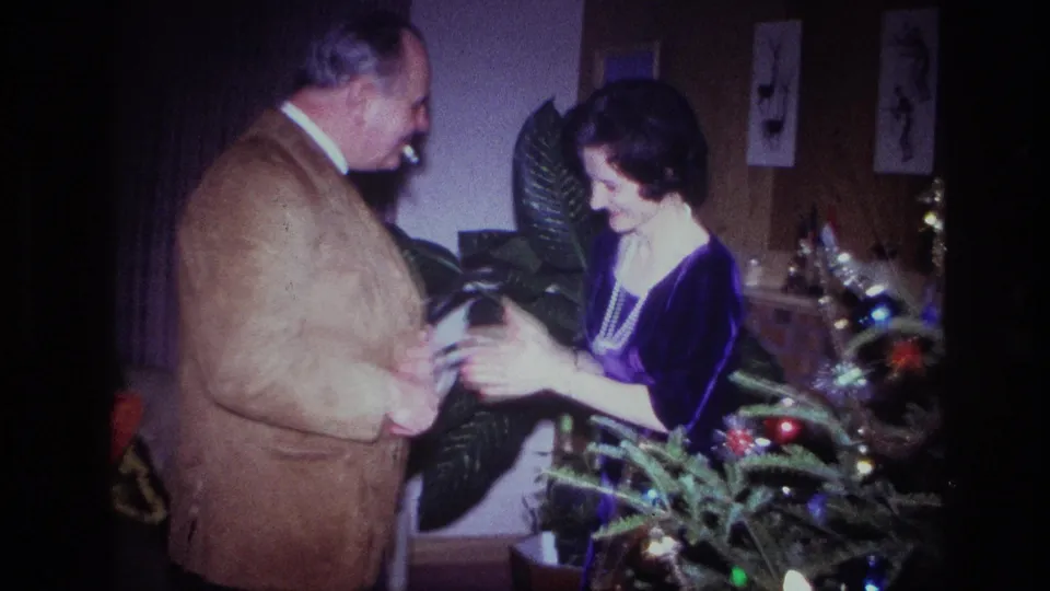 The woman gestures with both hands while the man faces her beside the lit Christmas tree in 1967 Ottawa footage.