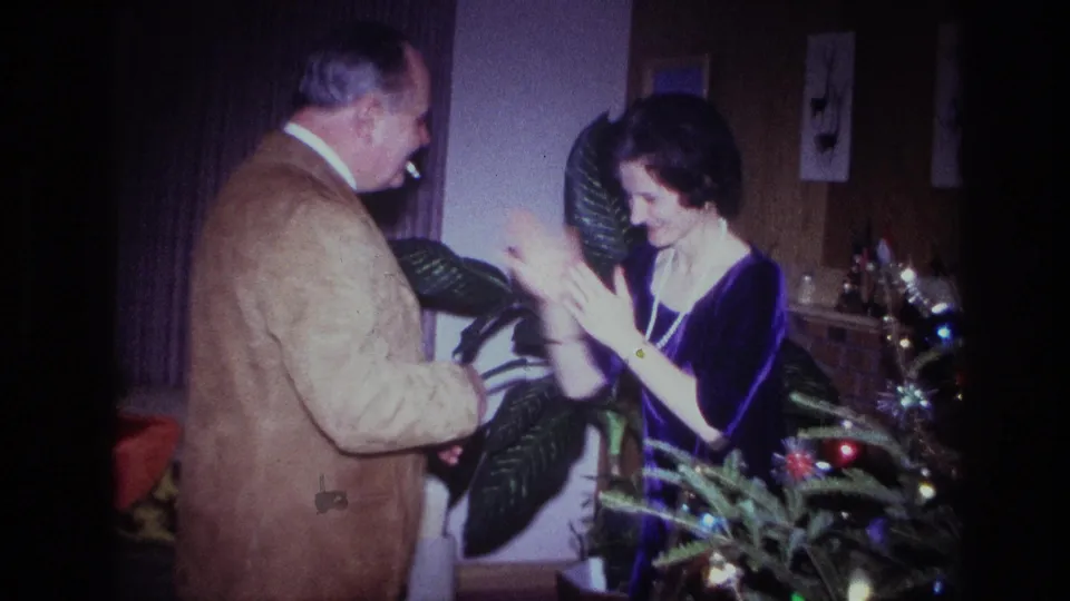 In a 1967 Ottawa home-movie frame, two adults smile and clap near houseplants and a small decorated Christmas tree.