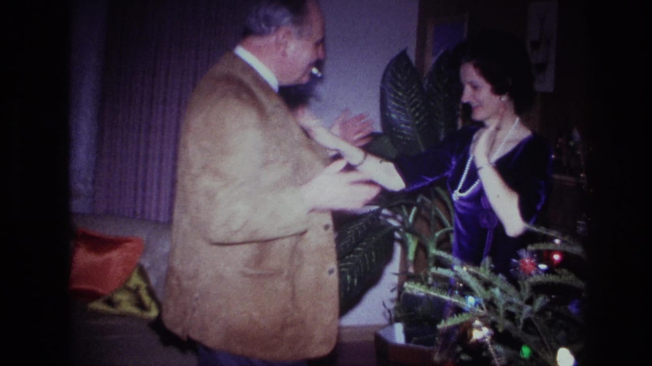Man reaches toward woman as they smile near the Christmas tree and houseplants.