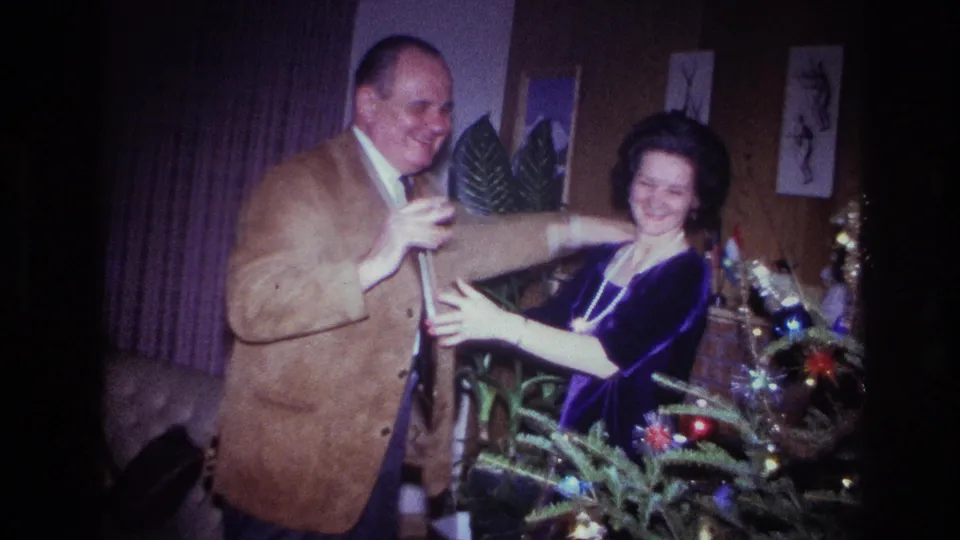 The man smiles while holding the woman's arm as they dance in a decorated 1967 Ottawa living room at Christmas.
