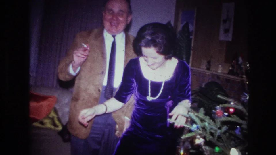 The couple laughs and gestures beside a small decorated tree in the Ottawa Christmas home movie, with indoor furniture behind them.