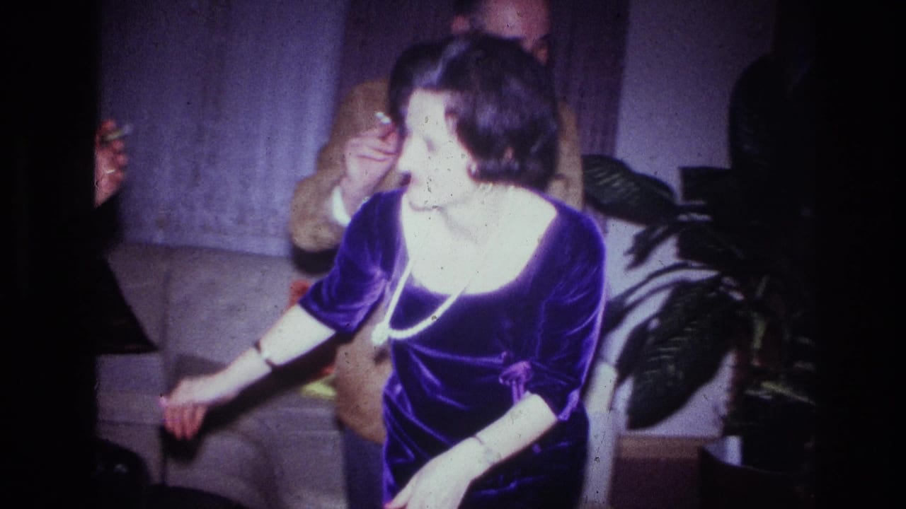 In the supplied 1967 Ottawa home-movie context, a woman in a purple dress and pearl necklace turns beside a sofa in a dim private interior.