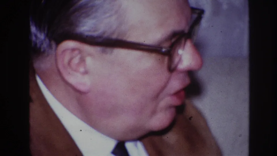 In the supplied 1967 Ottawa home-movie context, the man in glasses remains framed tightly in profile during the indoor party conversation.