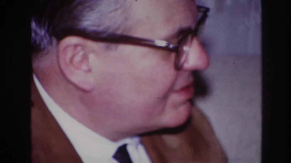 In the supplied 1967 Ottawa home-movie context, the close-up holds on the man in glasses and brown suit as he faces right during conversation.