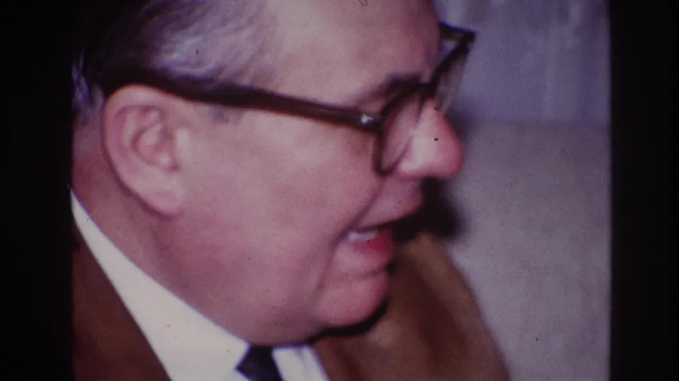 The 1967 Ottawa home-movie frame shows a suited man in glasses in side profile, apparently mid-sentence indoors.