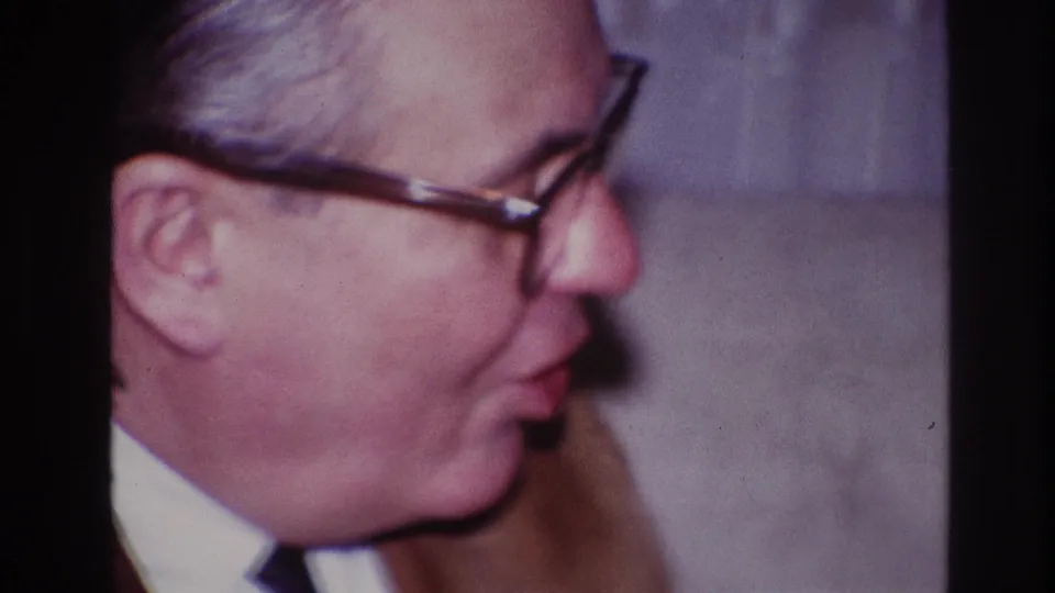 A close 8mm frame shows the gray-haired man with glasses in profile against a plain wall in the Ottawa interior scene.
