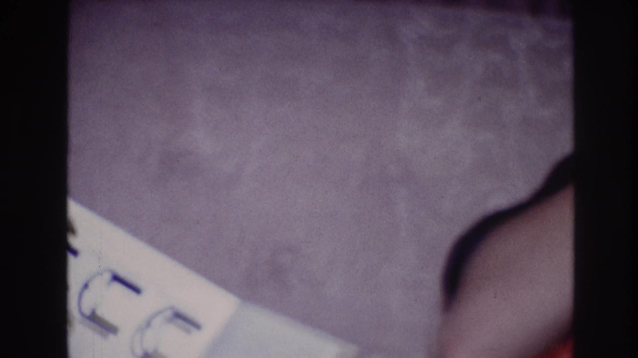 1967 Ottawa home-movie frame showing a partial wrist with a dark bracelet or watch near a white paper on purple carpet.