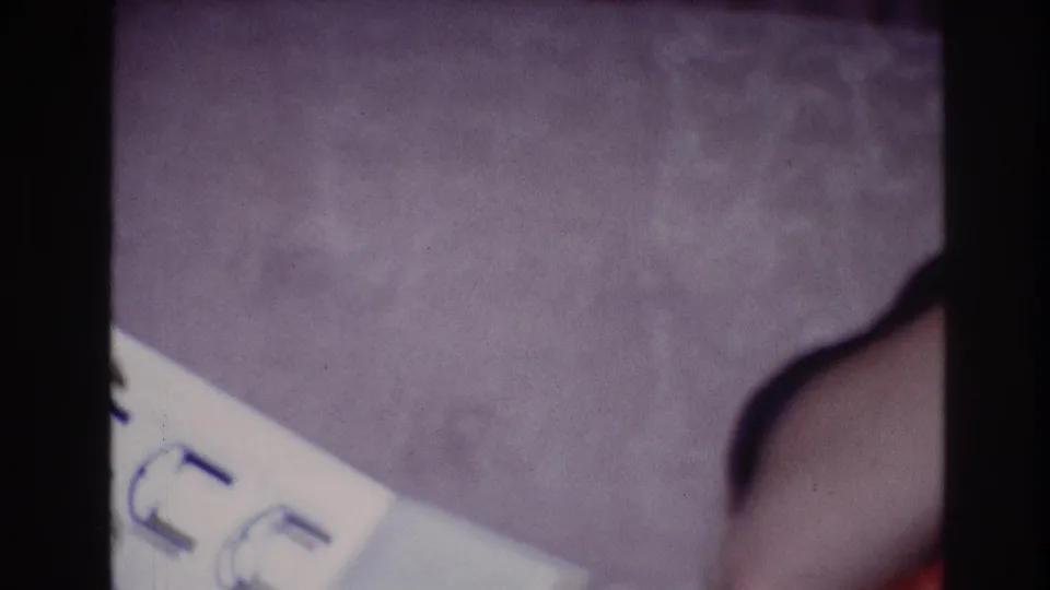 1967 Ottawa home-movie frame showing a partial wrist with a dark bracelet or watch near a white paper on purple carpet.