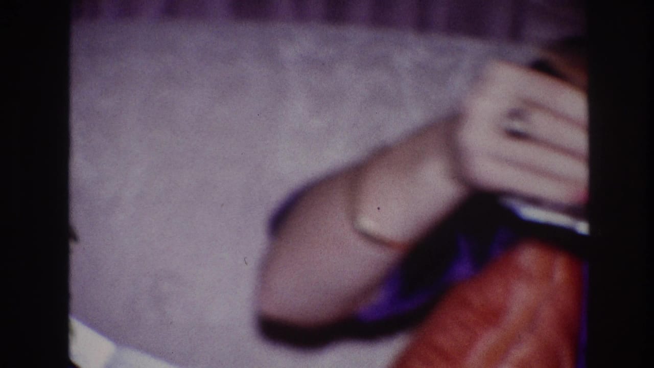 1967 Ottawa home-movie frame showing a woman’s arm and hand holding a drink near a light sofa, with a dark contact-sheet border.