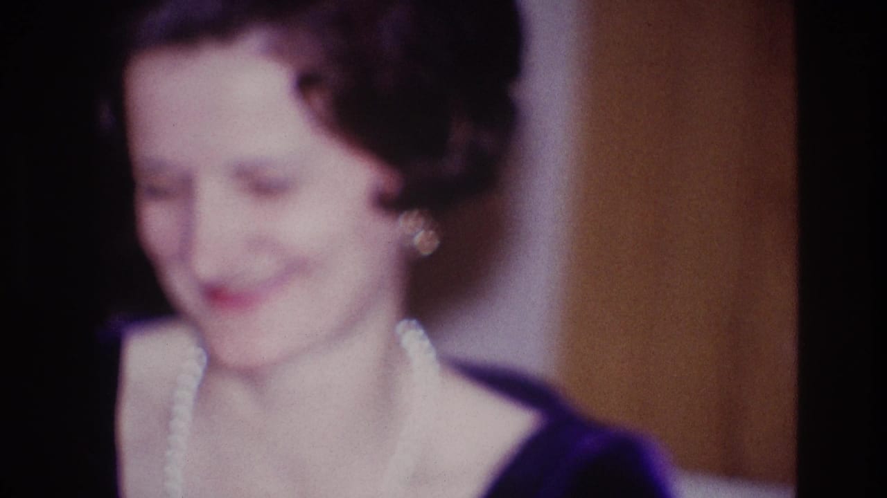 Smiling woman in 1967 Ottawa home-movie footage, wearing a dark dress, earrings and pearls in a warm private interior.