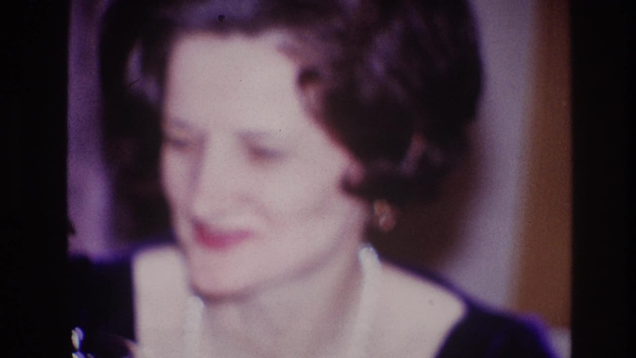 Older woman with dark curled hair smiles in soft focus inside a dim room.