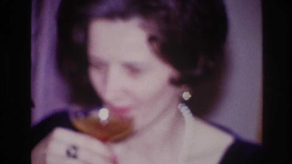 In the Ottawa 1967 home-movie frame, the woman raises a stemmed drink glass, with her ring and earrings catching light.