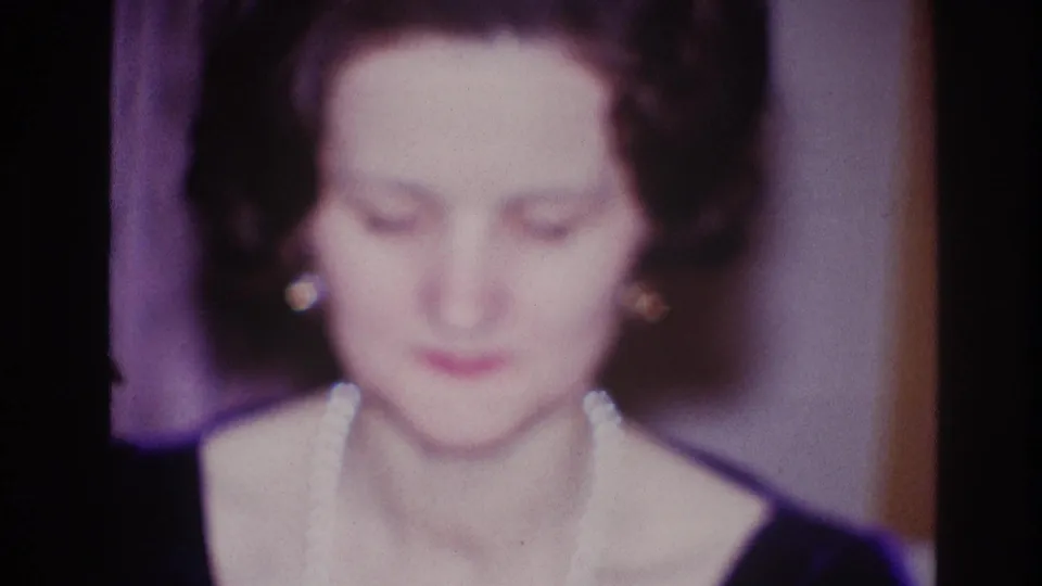 In 1967 Ottawa home-movie footage, a woman in a dark dress looks down beside curtains and a warm-toned interior wall.