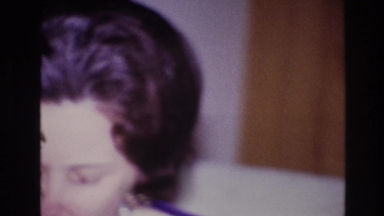The home-movie camera catches the woman's hair and lowered face at the edge of frame, with an interior wall behind her.