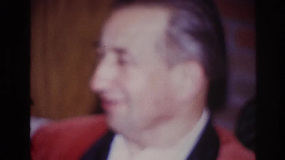 The man appears to smile in profile while seated on a couch in warm 8mm home-movie footage.