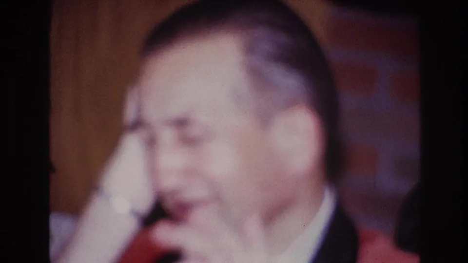 The seated man raises a hand to his head while wearing a red jacket in the 1967 Ottawa home-movie scene.