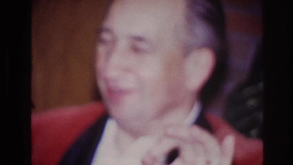 1967 Ottawa 8mm frame showing an adult man in a red jacket, seated indoors with his hands raised near his chin.