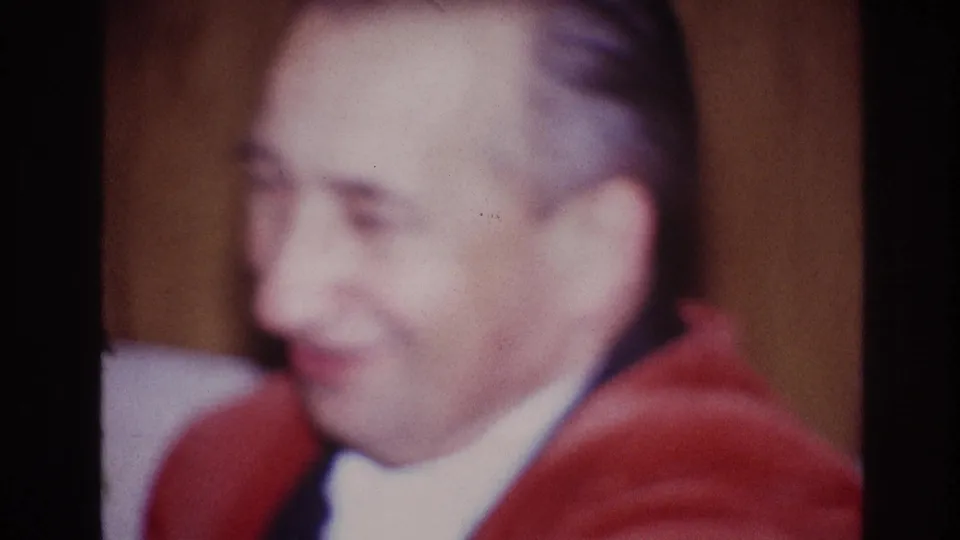 1967 Ottawa home-movie frame of an adult man in a red jacket looking down or to the side in a warm interior.