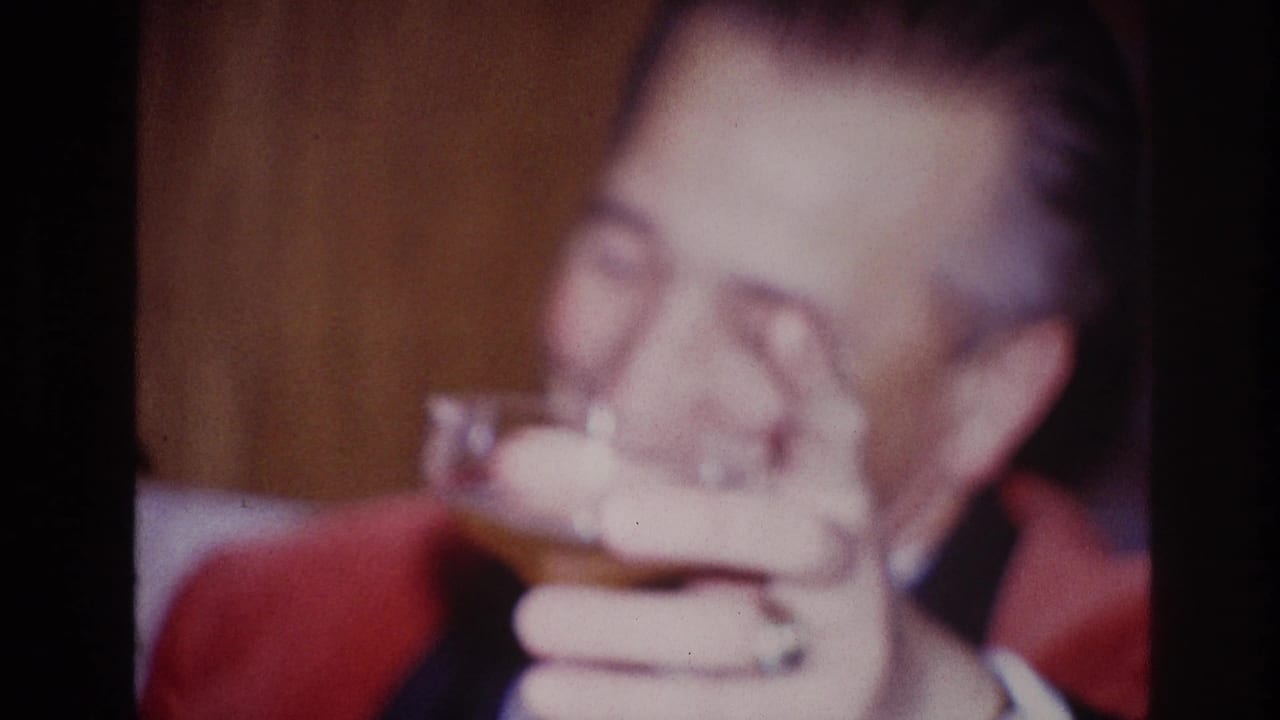 Older man in a red jacket lifting a small glass toward his mouth.