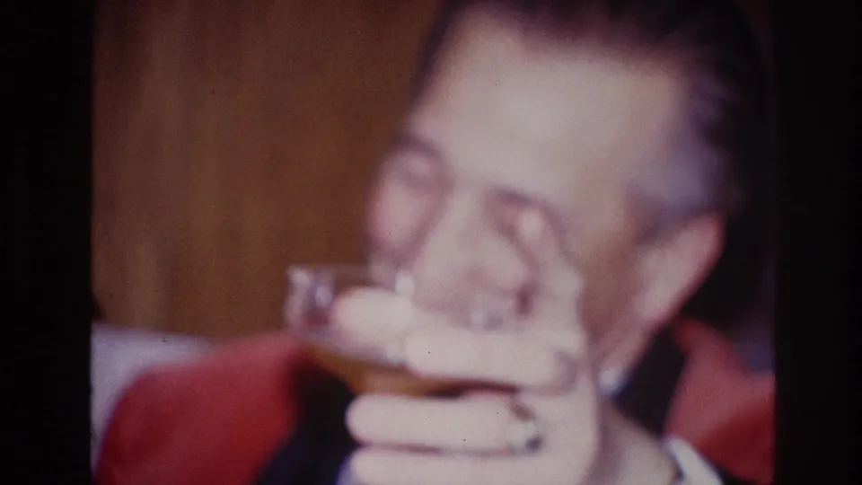 The older man raises a cocktail glass close to his face in a warmly lit Ottawa interior, smiling in soft 8mm focus.