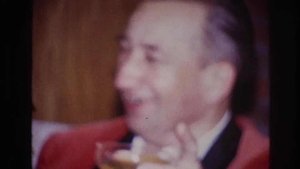 An older man in a red jacket and white shirt smiles in a private Ottawa interior as his drink drops lower in frame.