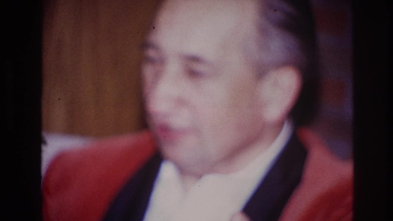 In 1967 Ottawa home-movie footage, an adult man in a red jacket and white shirt sits indoors in soft focus.