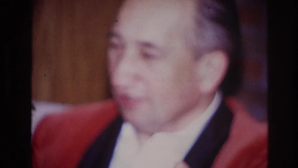 In 1967 Ottawa home-movie footage, an adult man in a red jacket and white shirt sits indoors in soft focus.