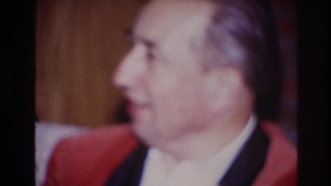 Adult man in red jacket turns in profile while seated indoors.