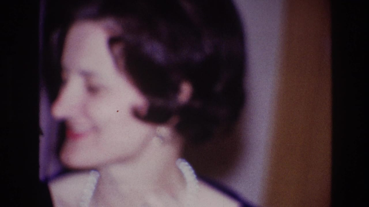 Profile view of the smiling woman in dark formal attire during the Ottawa 1967 home-movie clip, framed closely against an indoor backdrop.