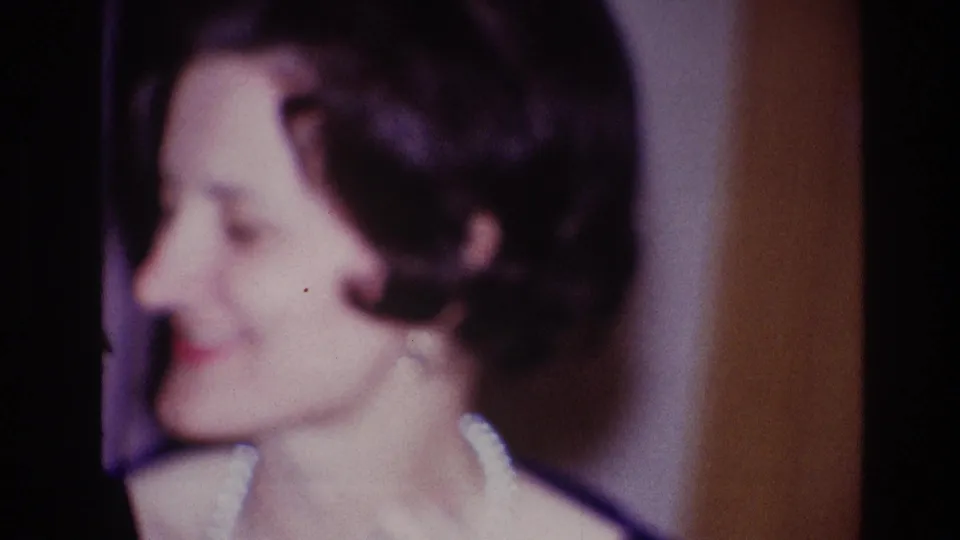 Profile view of the smiling woman in dark formal attire during the Ottawa 1967 home-movie clip, framed closely against an indoor backdrop.