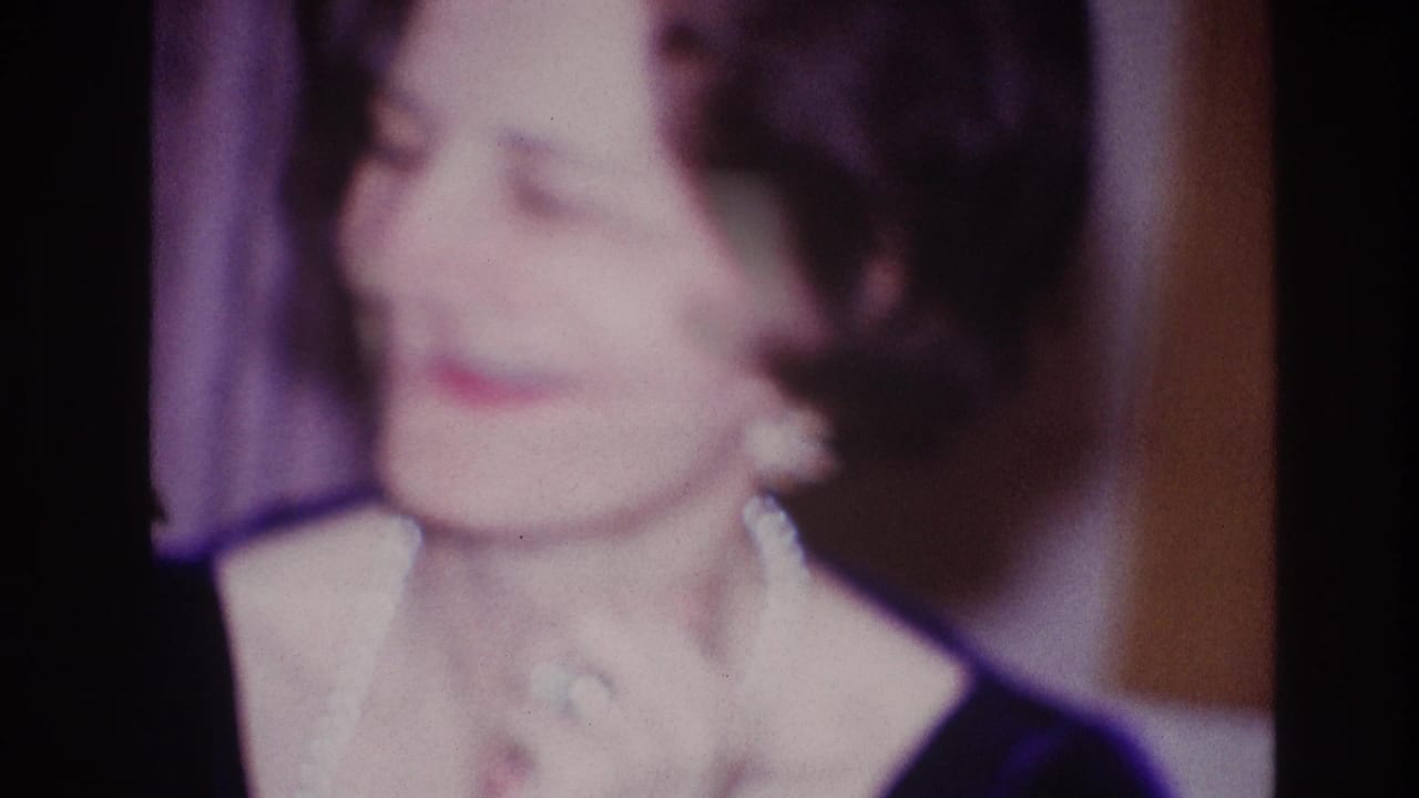 Adult woman in a dark dress and light necklace smiles while clapping in the Ottawa 1967 formal-event home-movie footage.