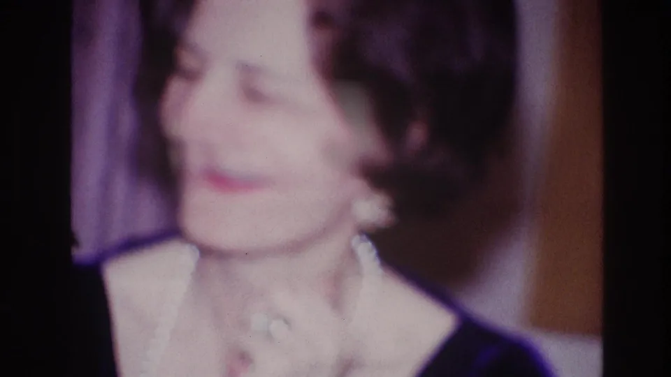 Adult woman in a dark dress and light necklace smiles while clapping in the Ottawa 1967 formal-event home-movie footage.