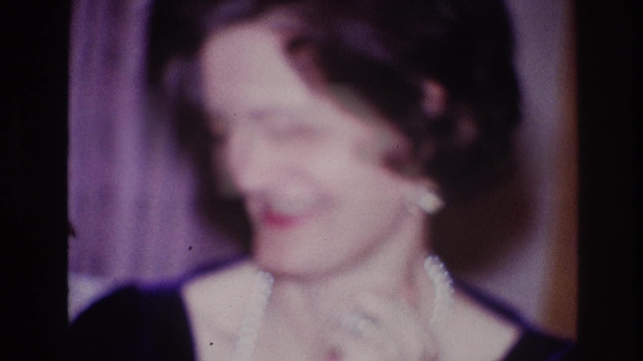 Soft-focus woman in a dark dress and pearl necklace looking down on a stage.