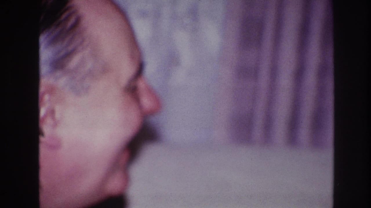 Older man in side profile smiling indoors beside a pale curtain.