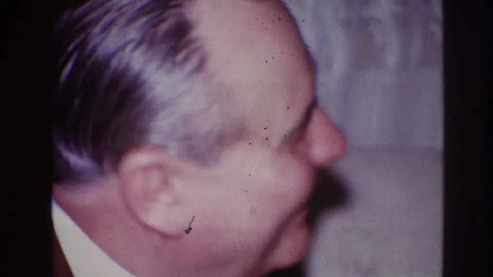 The 1967 Ottawa home-movie frame holds on the older man's smiling profile, with film grain and a pale curtain behind him.