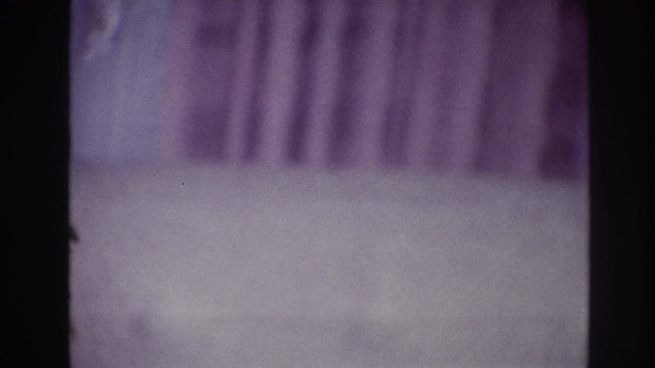 Ottawa 1967 home-movie frame with a purple curtain backdrop and soft interior detail, before the woman is clearly framed.