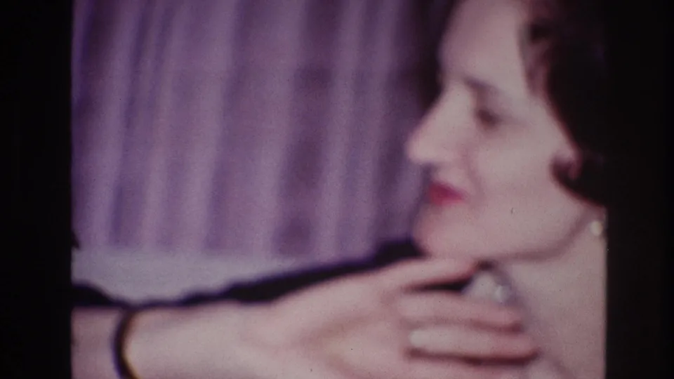 Adult woman in Ottawa 1967 home-movie footage, seen in soft profile while touching her neck near a purple curtain backdrop.