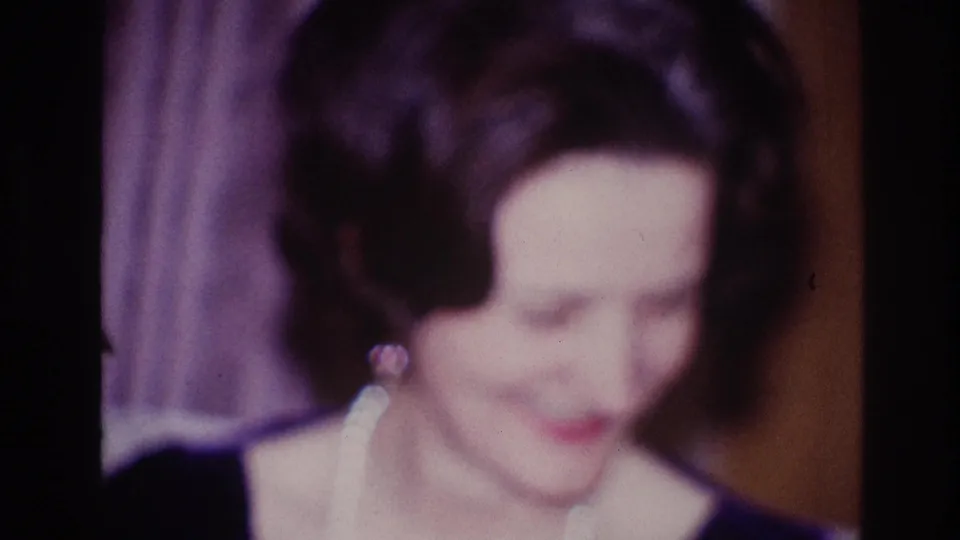 Adult woman in Ottawa 1967 home-movie footage, wearing a dark dress and pearl necklace while smiling downward in a soft close-up.