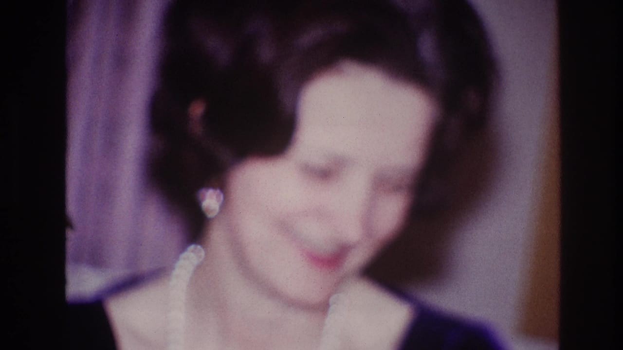 1967 Ottawa home-movie frame of a woman in a dark dress and pearls looking downward in a softly lit private interior.