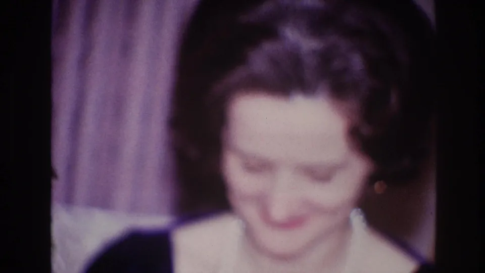 1967 Ottawa home-movie frame of the woman looking down, with pearls, earrings, and curtain backdrop visible.