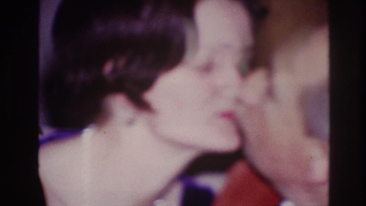 1967 Ottawa home-movie frame with a woman leaning toward a man in a red shirt for a kiss indoors.
