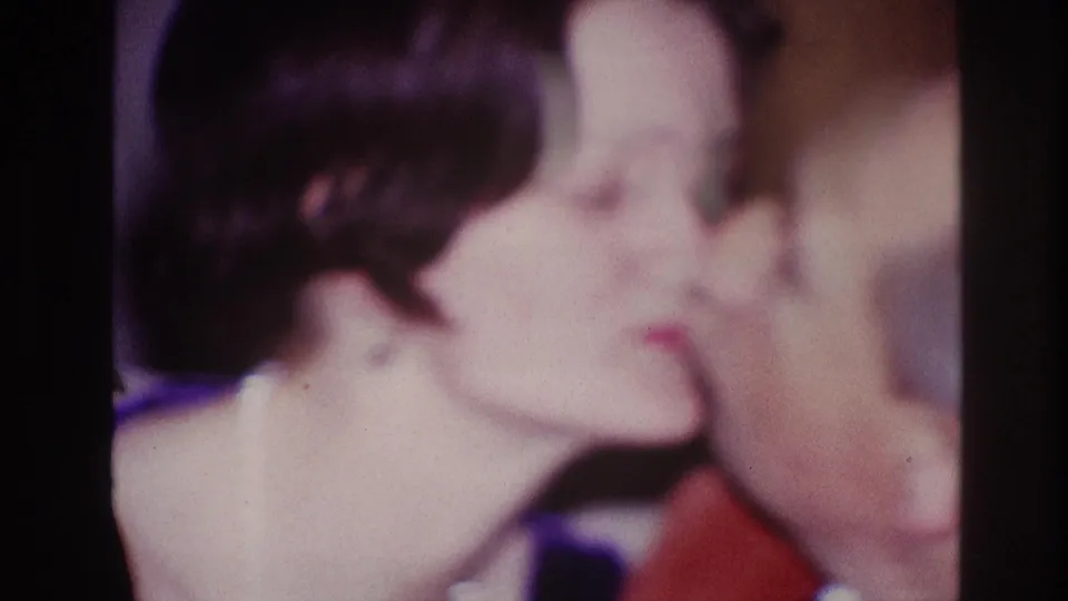 1967 Ottawa home-movie frame with a woman leaning toward a man in a red shirt for a kiss indoors.