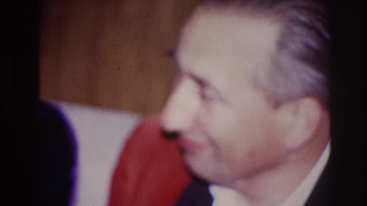 Seated man in a dark suit smiles in profile beside a woman in red.