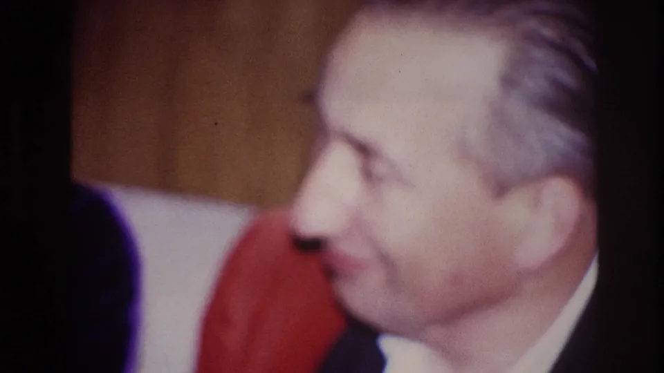 1967 Ottawa home-movie frame of a seated man in a dark suit smiling beside a woman wearing red in a private interior.