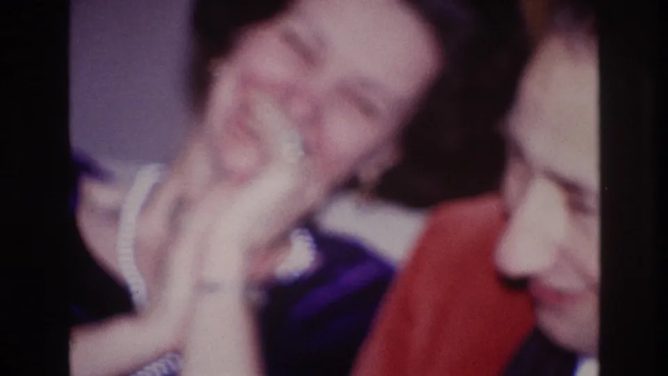 1967 Ottawa home-movie frame of a woman laughing with her hand raised while seated beside a suited man.