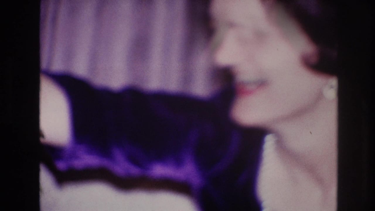 In this 1967 Ottawa home-movie frame, a woman in a dark velvet dress and pearl necklace smiles in profile near a curtain backdrop.